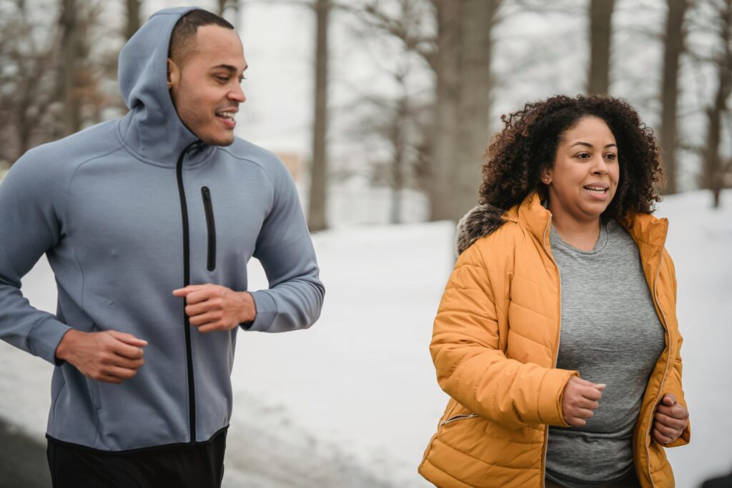 Positive African American male personal instructor running with overweight female in park in winter day