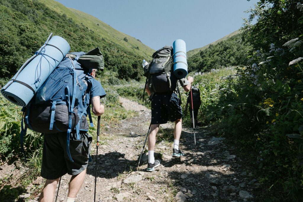 Two backpackers ascending a sunny, rocky mountain path with trekking poles in hand.
