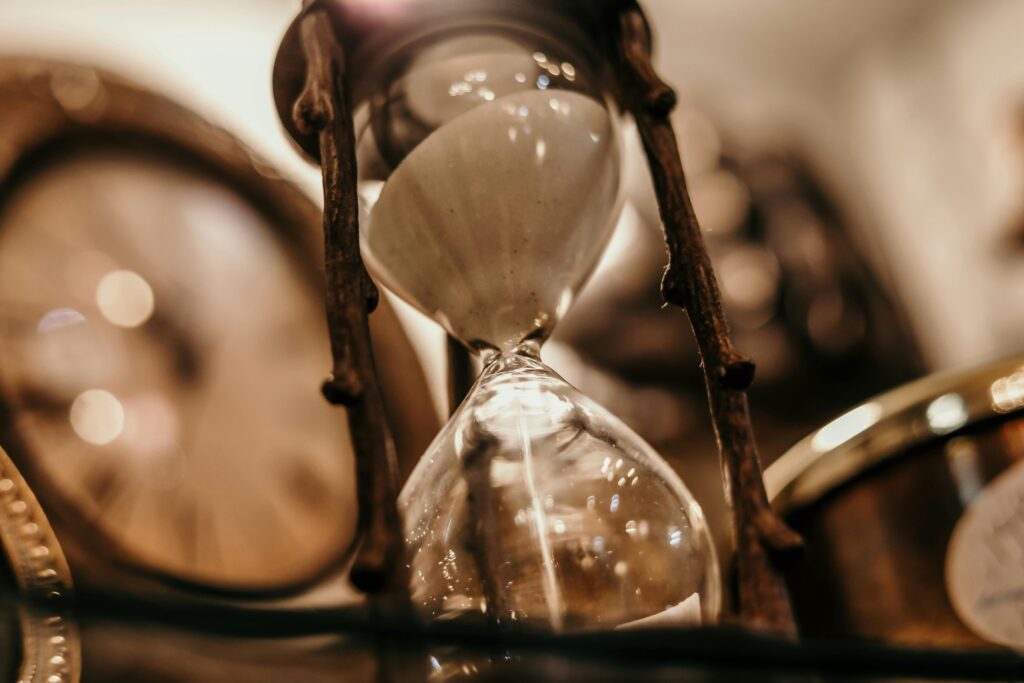 Close-up of a vintage hourglass with blurred antique clock background, evoking a timeless feel.